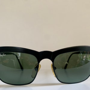 May include: Black sunglasses with a matte finish and green lenses. The Ray-Ban logo is visible on the left lens. The sunglasses have a classic, semi-rimless design.