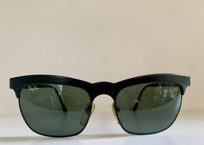 May include: Black sunglasses with a matte finish and green lenses. The Ray-Ban logo is visible on the left lens. The sunglasses have a classic, semi-rimless design.