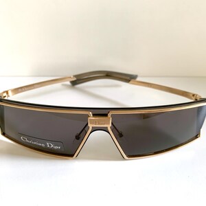 Peut inclure: Lunettes de soleil Christian Dior avec une monture dor&eacute;e et des verres gris fonc&eacute;. Les lunettes ont une forme rectangulaire avec un d&eacute;tail de pont et le nom de la marque sur la monture. Les branches sont dor&eacute;es.