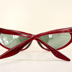 May include: Red glitter sunglasses with a cat-eye frame. The Ray-Ban logo is visible on the left temple. The lenses are a muted green color. The sunglasses are angled, showing the shape of the frame and lenses.