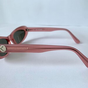 Peut inclure: Lunettes de soleil roses en forme d'&oelig;il de chat avec des verres fonc&eacute;s. Les montures sont de couleur rose translucide et les branches ont un embl&egrave;me argent&eacute;. Les lunettes de soleil sont pr&eacute;sent&eacute;es de profil.