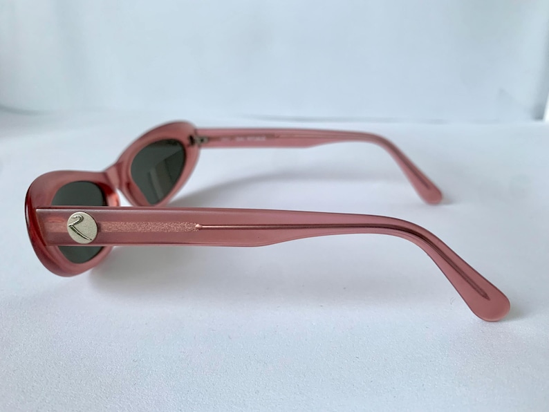 Peut inclure: Lunettes de soleil roses en forme d'&oelig;il de chat avec des verres fonc&eacute;s. Les montures sont de couleur rose translucide et les branches ont un embl&egrave;me argent&eacute;. Les lunettes de soleil sont pr&eacute;sent&eacute;es de profil.