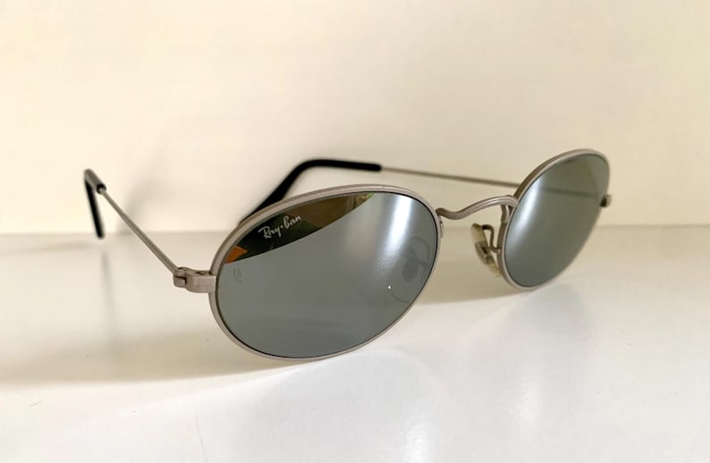Puede incluir: Gafas de sol Ray-Ban plateadas con lentes ovalados y montura plateada. Las gafas tienen un acabado espejado.