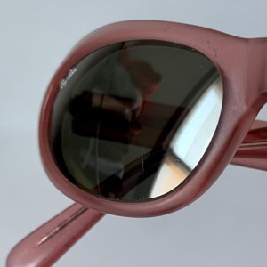 Peut inclure: Gros plan sur des lunettes de soleil roses avec des verres fonc&eacute;s. Les lunettes ont une monture arrondie et un logo subtil sur le verre. Les verres refl&egrave;tent l'environnement. Le design g&eacute;n&eacute;ral est r&eacute;tro et &eacute;l&eacute;gant.