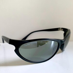 May include: Black sunglasses with a wrap-around frame design and dark tinted lenses. The left lens features the white Ray-Ban logo printed in the upper corner. The frames are constructed from a matte black material with curved temples.