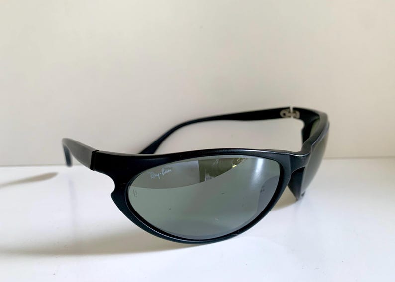 May include: Black sunglasses with a wrap-around frame design and dark tinted lenses. The left lens features the white Ray-Ban logo printed in the upper corner. The frames are constructed from a matte black material with curved temples.