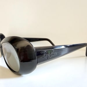 May include: Black Ray-Ban sunglasses with thick, rounded frames and grey lenses. The Ray-Ban logo is visible on the temple. The sunglasses are set against a white background.