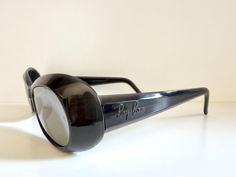 May include: Black Ray-Ban sunglasses with thick, rounded frames and grey lenses. The Ray-Ban logo is visible on the temple. The sunglasses are set against a white background.
