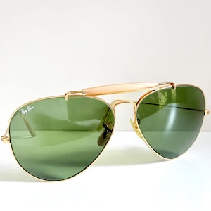 May include: Gold-framed aviator sunglasses with green lenses. The sunglasses feature a bridge bar and a light-colored brow bar. The Ray-Ban logo is visible on the lens. A classic eyewear accessory.