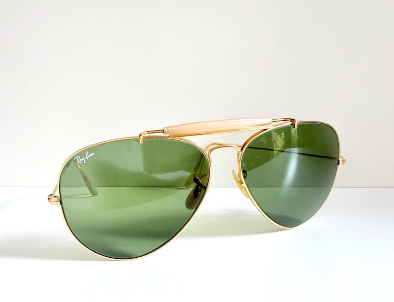 May include: Gold-framed aviator sunglasses with green lenses. The sunglasses feature a bridge bar and a light-colored brow bar. The Ray-Ban logo is visible on the lens. A classic eyewear accessory.