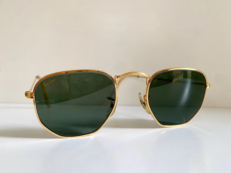 Może przedstawiać: Okulary przeciwsłoneczne w złotej oprawie z ciemnozielonymi soczewkami. Logo Ray-Ban jest widoczne na soczewce. Okulary przeciwsłoneczne mają klasyczny, geometryczny kształt i są przeznaczone do ochrony przed słońcem.