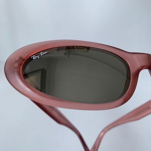 Peut inclure: Gros plan sur des lunettes de soleil Ray-Ban roses. Les lunettes ont une monture arrondie et des verres fonc&eacute;s. Le logo Ray-Ban est visible en haut &agrave; gauche de la monture. Les branches des lunettes sont fines et s'&eacute;tendent vers le bas.