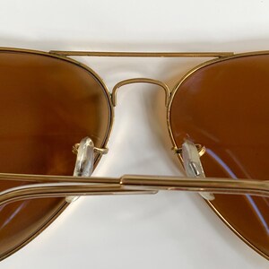 May include: Close-up of gold-framed aviator sunglasses with brown lenses. The sunglasses have a double bridge and clear nose pads. The lenses are a warm brown colour, and the frames have a shiny gold finish.