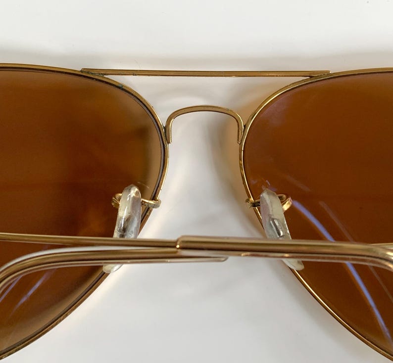 May include: Close-up of gold-framed aviator sunglasses with brown lenses. The sunglasses have a double bridge and clear nose pads. The lenses are a warm brown colour, and the frames have a shiny gold finish.