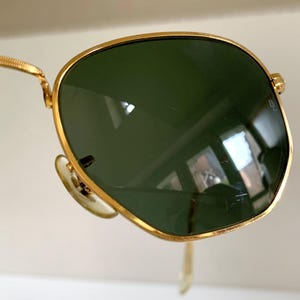 Może przedstawiać: Okulary przeciwsłoneczne ze złotą oprawką i zielonymi soczewkami. Okulary przeciwsłoneczne w stylu aviator mają złotą oprawkę i ciemnozielone soczewki. Elegancki i nowoczesny design, dla mężczyzn i kobiet.