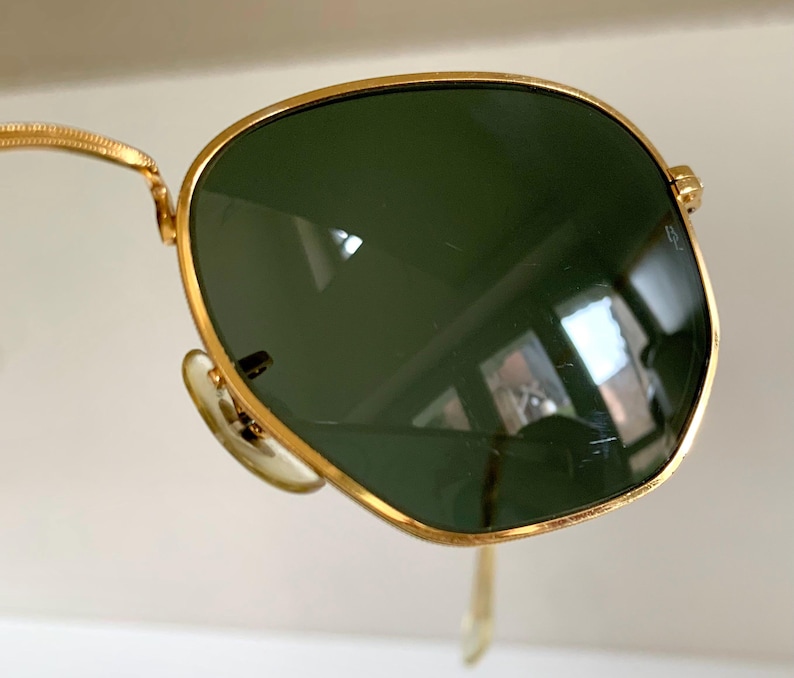 Może przedstawiać: Okulary przeciwsłoneczne ze złotą oprawką i zielonymi soczewkami. Okulary przeciwsłoneczne w stylu aviator mają złotą oprawkę i ciemnozielone soczewki. Elegancki i nowoczesny design, dla mężczyzn i kobiet.