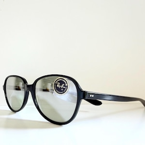 May include: Black oversized sunglasses with silver lenses. The sunglasses have a rounded frame and a sticker on the right lens. The brand name Ray-Ban is visible on the sticker.