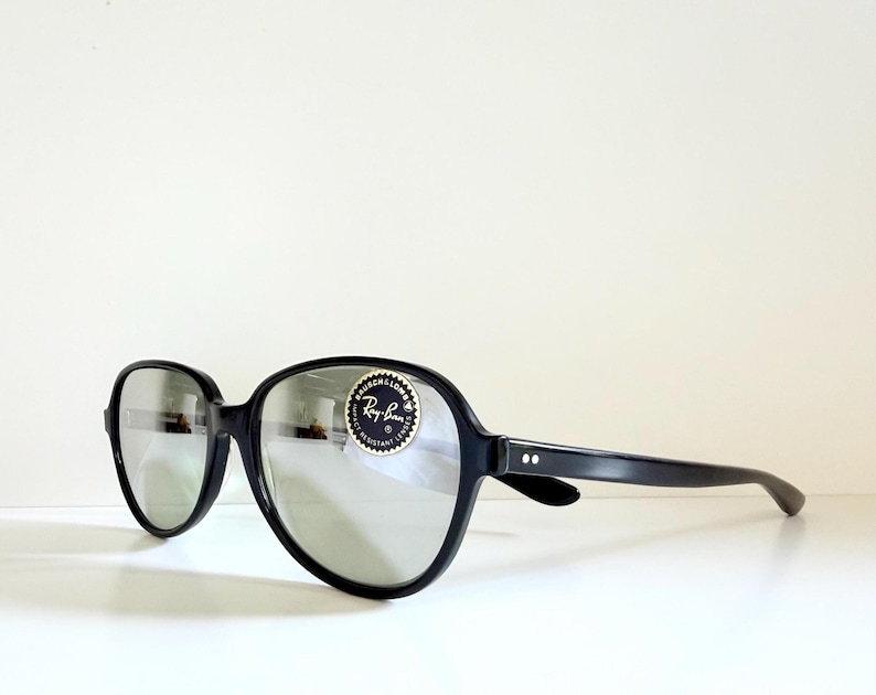 May include: Black oversized sunglasses with silver lenses. The sunglasses have a rounded frame and a sticker on the right lens. The brand name Ray-Ban is visible on the sticker.