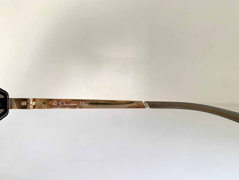 Peut inclure: Gros plan sur une branche de lunettes dor&eacute;e avec le logo Christian Dior. La branche passe &agrave; une couleur brun-gris plus fonc&eacute;e au niveau de la tempe. Le design pr&eacute;sente une charni&egrave;re et une esth&eacute;tique &eacute;l&eacute;gante et moderne.