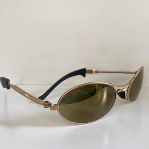 May include: Gold-tone oval sunglasses with brown lenses. The sunglasses have a sleek, modern design with black temple tips and a subtle Ray-Ban logo on the lens. The frames are a light gold color.