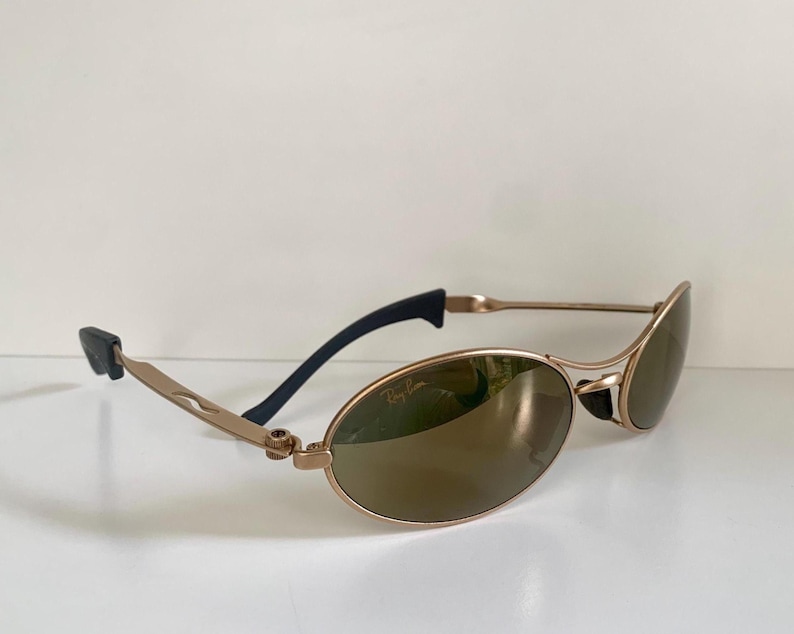 May include: Gold-tone oval sunglasses with brown lenses. The sunglasses have a sleek, modern design with black temple tips and a subtle Ray-Ban logo on the lens. The frames are a light gold color.