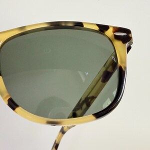 May include: Close-up of sunglasses with a tortoiseshell pattern frame and dark green lenses. The sunglasses have a classic design with a rectangular shape. The frame features a mix of light brown, dark brown, and black spots.