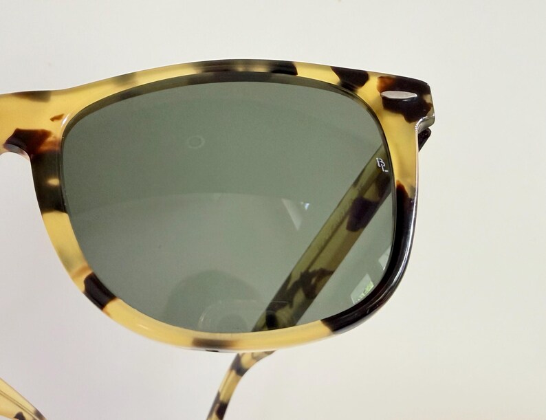 May include: Close-up of sunglasses with a tortoiseshell pattern frame and dark green lenses. The sunglasses have a classic design with a rectangular shape. The frame features a mix of light brown, dark brown, and black spots.