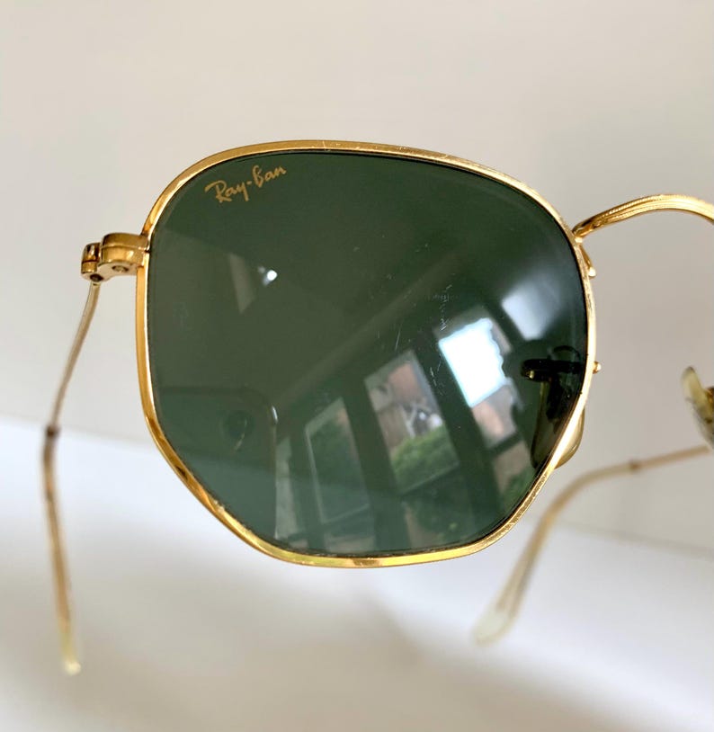 Może przedstawiać: Zbliżenie okular&oacute;w przeciwsłonecznych ze złotą oprawką i zielonymi soczewkami. Logo Ray-Ban jest widoczne na g&oacute;rnej lewej soczewce. Okulary mają klasyczny, prostokątny kształt z cienkimi, złotymi zausznikami.