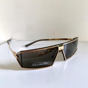Peut inclure: Lunettes de soleil &agrave; monture dor&eacute;e avec verres fonc&eacute;s. Les lunettes ont une forme rectangulaire avec un pont et des branches dor&eacute;s. Le texte "Christian Dior" est visible sur le verre.