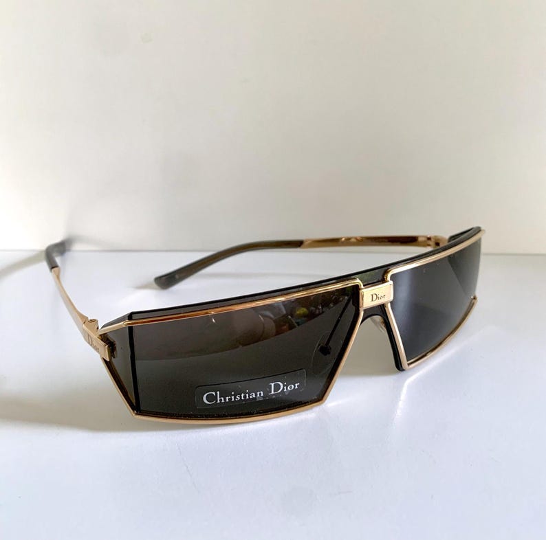 Peut inclure: Lunettes de soleil &agrave; monture dor&eacute;e avec verres fonc&eacute;s. Les lunettes ont une forme rectangulaire avec un pont et des branches dor&eacute;s. Le texte "Christian Dior" est visible sur le verre.