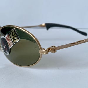 May include: Gold-tone sunglasses with oval lenses and a black arm. The glasses have a sticker that says "SPORT" and features the Olympic rings. The frame is a light gold color.