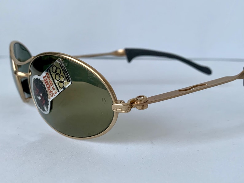 May include: Gold-tone sunglasses with oval lenses and a black arm. The glasses have a sticker that says "SPORT" and features the Olympic rings. The frame is a light gold color.