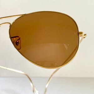May include: Gold-framed aviator sunglasses with brown lenses. The lenses have a teardrop shape, and the frames are thin and delicate. The sunglasses are set against a white background.
