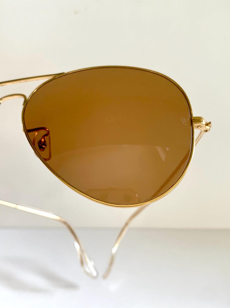 May include: Gold-framed aviator sunglasses with brown lenses. The lenses have a teardrop shape, and the frames are thin and delicate. The sunglasses are set against a white background.