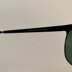 May include: Close-up of black sunglasses with green lenses. The text "B&L RAY-BAN/FRAME FRANCE" is printed on the temple arm. The sunglasses have a classic, rectangular shape.