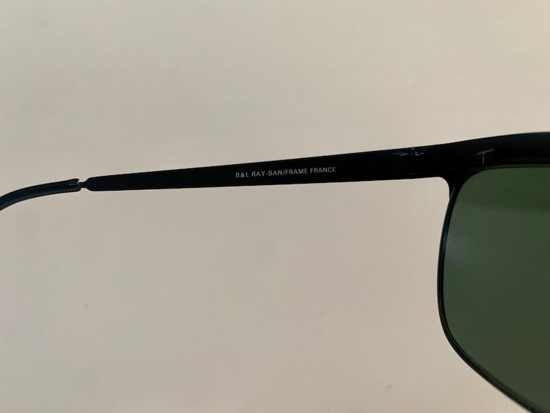 May include: Close-up of black sunglasses with green lenses. The text "B&L RAY-BAN/FRAME FRANCE" is printed on the temple arm. The sunglasses have a classic, rectangular shape.