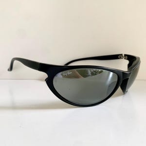 May include: Black sunglasses with a wraparound frame design and dark, reflective lenses. The right lens features the white Ray-Ban logo printed in the upper corner. The temples are solid black and curve slightly at the ends for a secure fit.