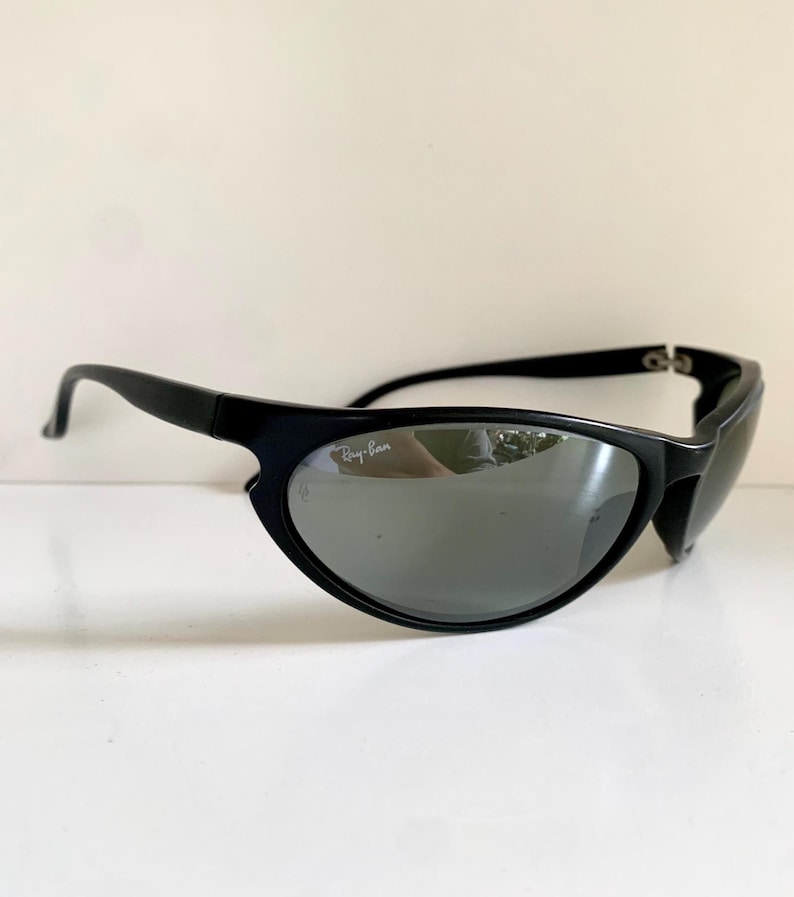 May include: Black sunglasses with a wraparound frame design and dark, reflective lenses. The right lens features the white Ray-Ban logo printed in the upper corner. The temples are solid black and curve slightly at the ends for a secure fit.