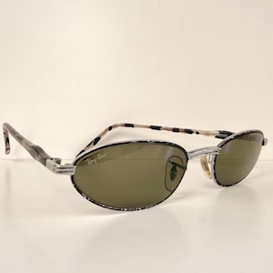 May include: Oval sunglasses with a black and white patterned frame and silver metal detailing. The lenses are a muted green, and the Ray-Ban logo is visible on the right lens.