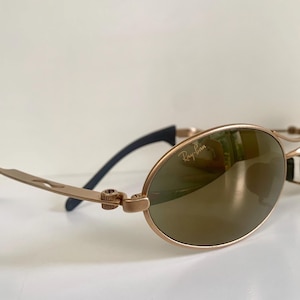 May include: Gold-toned Ray-Ban sunglasses with oval lenses and a sleek, modern design. The lenses have a reflective, bronze tint. The temples are a light gold color with black accents near the ear pieces. The Ray-Ban logo is visible on the lens.