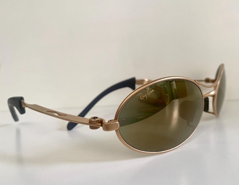 May include: Gold-toned Ray-Ban sunglasses with oval lenses and a sleek, modern design. The lenses have a reflective, bronze tint. The temples are a light gold color with black accents near the ear pieces. The Ray-Ban logo is visible on the lens.