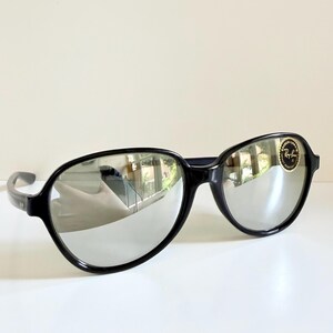May include: Black sunglasses with mirrored lenses. The sunglasses have a rounded frame and a small gold and black logo sticker on the right lens. The lenses reflect the surrounding environment.