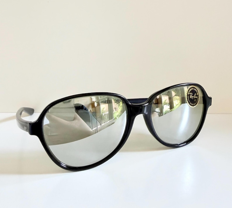 May include: Black sunglasses with mirrored lenses. The sunglasses have a rounded frame and a small gold and black logo sticker on the right lens. The lenses reflect the surrounding environment.