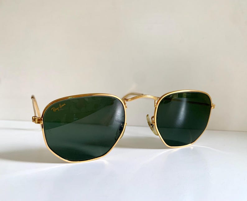 Może przedstawiać: Okulary przeciwsłoneczne w złotej oprawie z ciemnozielonymi soczewkami. Okulary mają charakterystyczny sześciokątny kształt, a logo Ray-Ban jest widoczne na soczewce. Oprawki są w błyszczącym złotym kolorze.