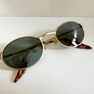 Puede incluir: Gafas de sol ovaladas con montura dorada y lentes verde oscuro. Las patillas presentan un dise&ntilde;o de carey. Las gafas de sol est&aacute;n sobre una superficie blanca.