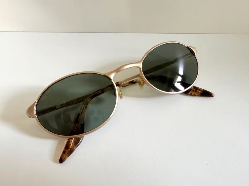 Puede incluir: Gafas de sol ovaladas con montura dorada y lentes verde oscuro. Las patillas presentan un dise&ntilde;o de carey. Las gafas de sol est&aacute;n sobre una superficie blanca.