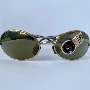 May include: Gold-framed sunglasses with oval lenses and a dark tint. The sunglasses have a black bridge and temples. A sticker on the right lens reads "SPORT" and features the Olympic rings. The Ray-Ban logo is visible on the left lens.
