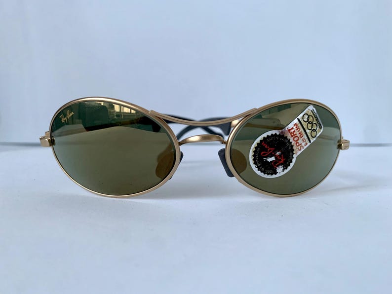 May include: Gold-framed sunglasses with oval lenses and a dark tint. The sunglasses have a black bridge and temples. A sticker on the right lens reads "SPORT" and features the Olympic rings. The Ray-Ban logo is visible on the left lens.