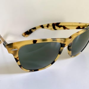 May include: Tortoiseshell sunglasses with dark lenses. The frames have a classic design with a light brown and black pattern. The sunglasses are angled, showing the shape of the lenses and the arms.