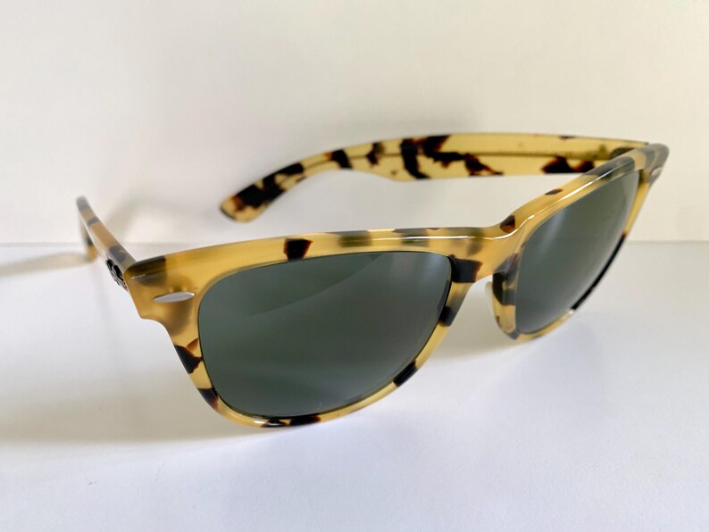 May include: Tortoiseshell sunglasses with dark lenses. The frames have a classic design with a light brown and black pattern. The sunglasses are angled, showing the shape of the lenses and the arms.